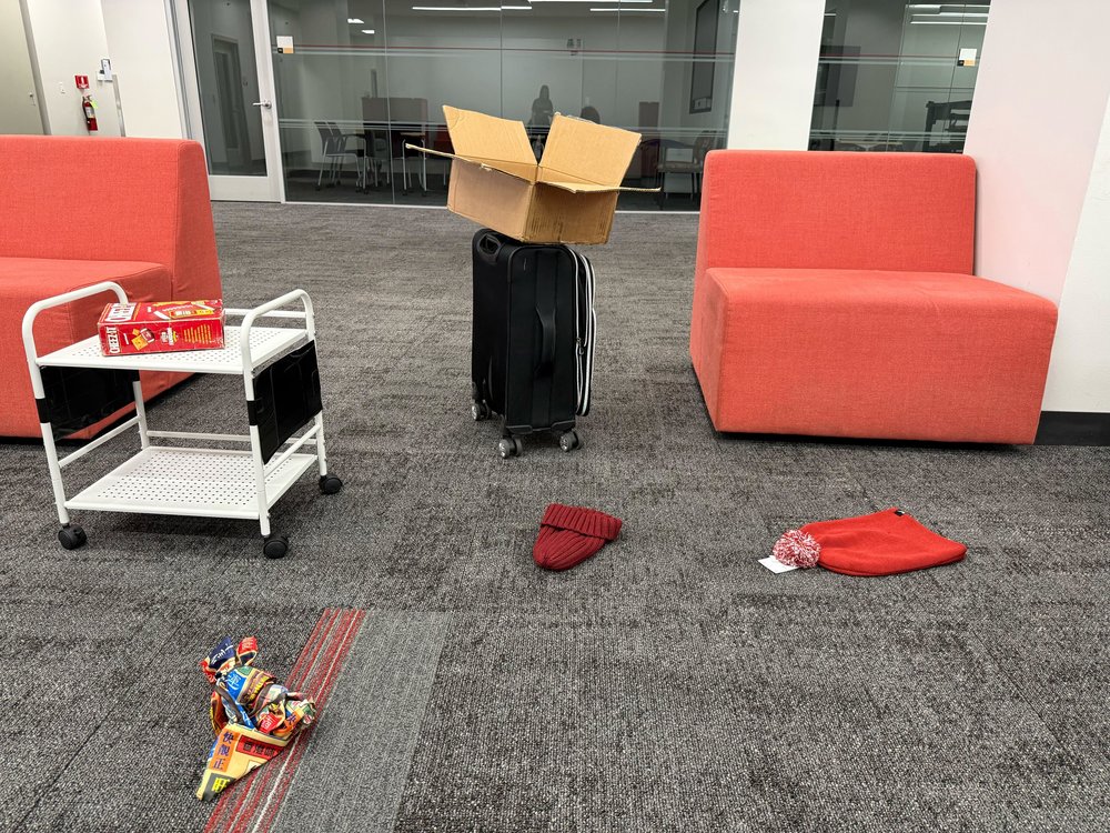 Conference room with a white rolling cart, a black suitcase topped with a cardboard box, red sofas, hats and a wrapped package on the floor